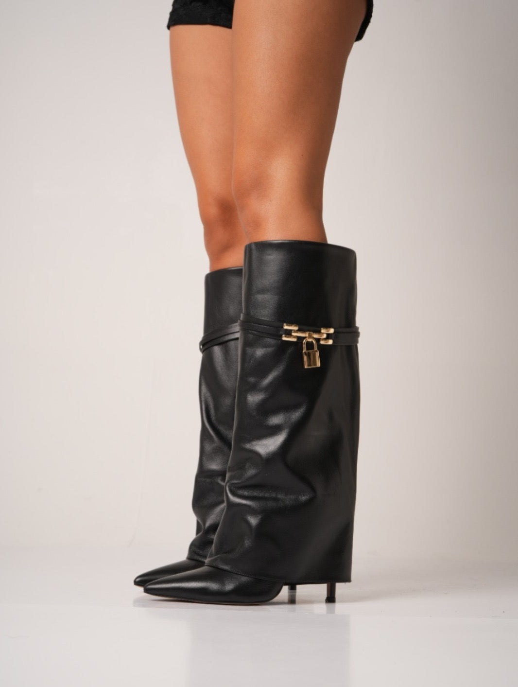 Elegant black knee-high stiletto boots with pointed toes, slouchy leather texture, and a gold padlock detail at the ankle. These statement boots blend luxury and edge—perfect for high-fashion looks or a bold night out.