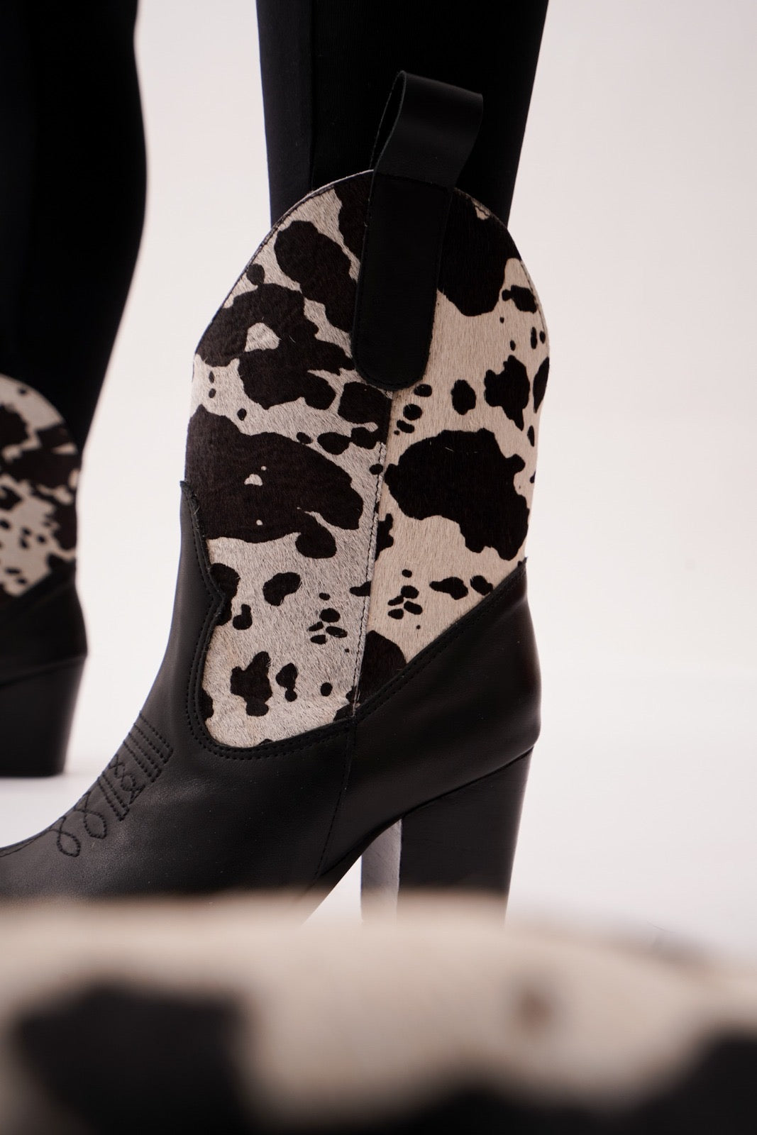 Black leather ankle boots featuring warm black-and-white cow-print uppers, wooden stacked heels, and engraved detailing. A bold Western statement for stylish day-to-night wear.