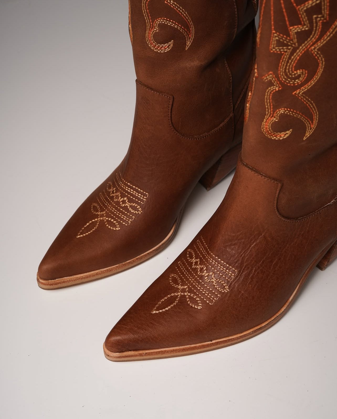 Tall tan cowboy boots with pointed toes, angled heels, and subtle Western embroidery along the shaft. A sleek and edgy twist on a classic silhouette, perfect for bold, versatile styling.