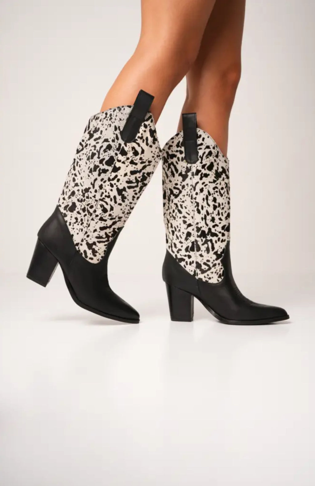 Black and white cow-print cowboy boots with high block heels, pointed toes, and pull tabs. These bold Western-style boots blend modern flair with ranch-inspired fashion, perfect for standout streetwear looks.