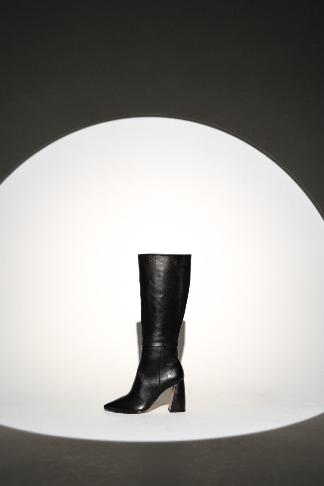 Sleek black knee-high leather boot with a pointed toe and sturdy high block heel. A minimalist and elegant design perfect for chic, everyday wear or polished evening looks.