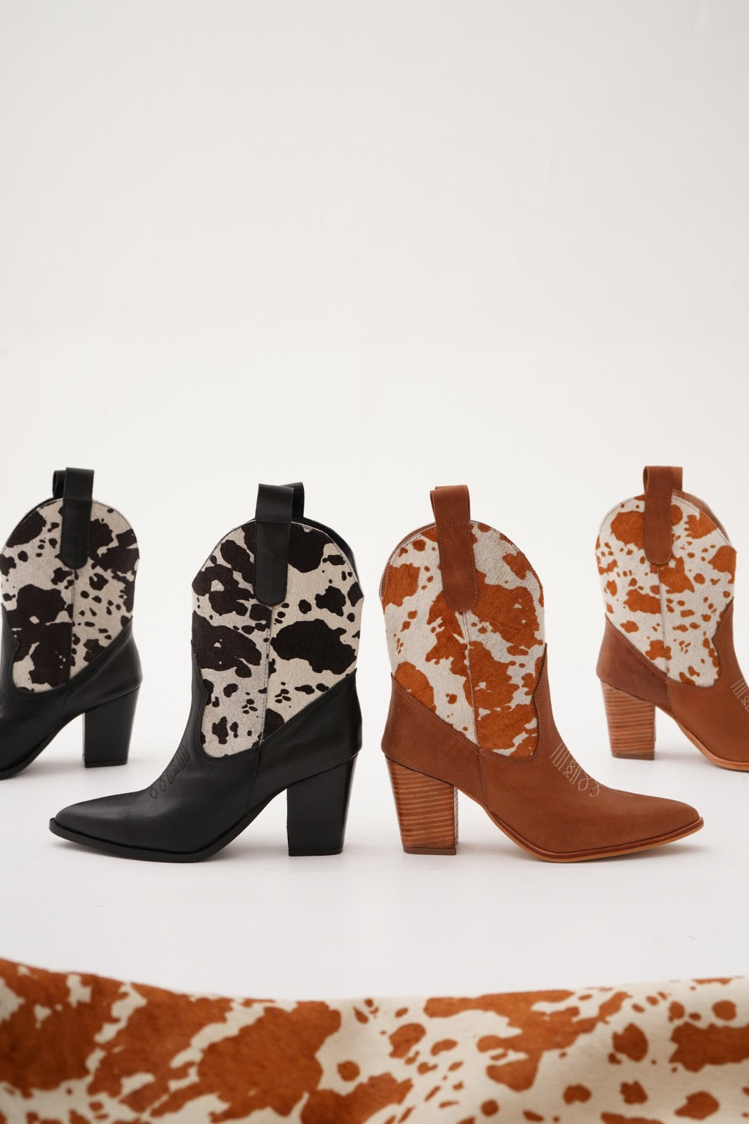 Rich black and tan leather ankle boots featuring warm orange-and-white cow-print uppers, wooden stacked heels, and engraved detailing. A bold Western statement for stylish day-to-night wear.
