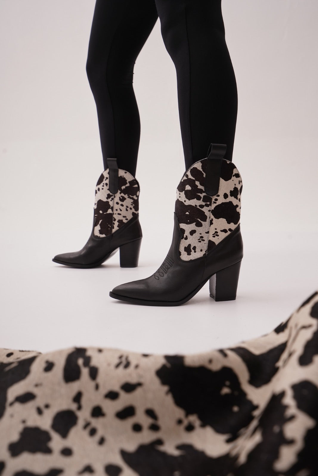 Black leather ankle boots featuring warm black-and-white cow-print uppers, wooden stacked heels, and engraved detailing. A bold Western statement for stylish day-to-night wear.