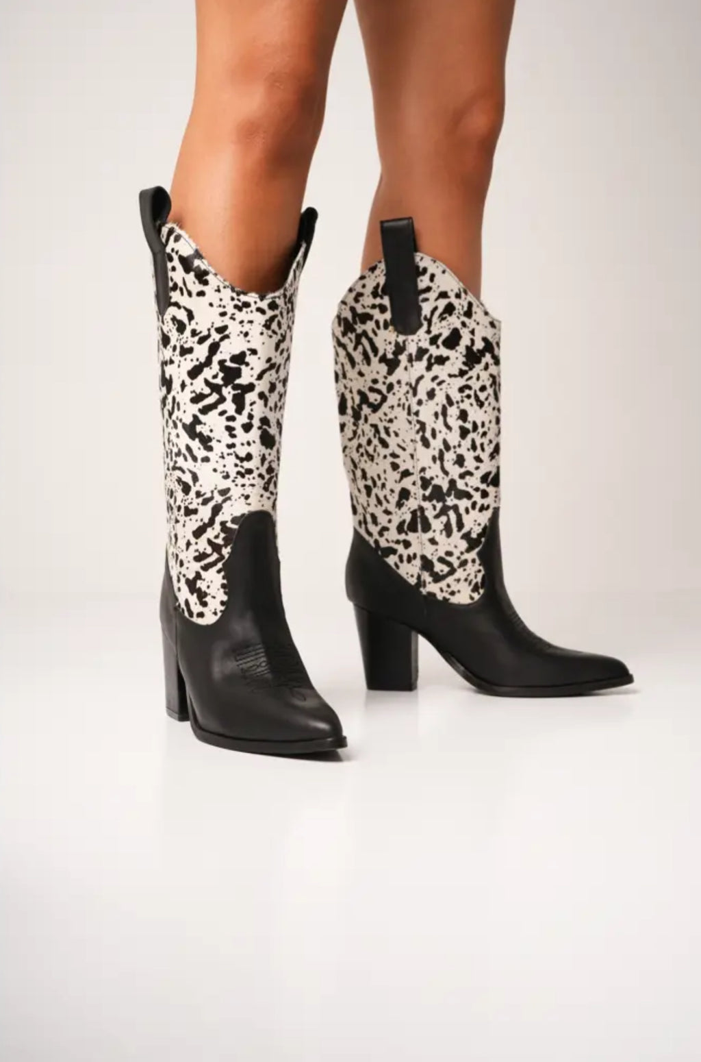 Black and white cow-print cowboy boots with high block heels, pointed toes, and pull tabs. These bold Western-style boots blend modern flair with ranch-inspired fashion, perfect for standout streetwear looks.