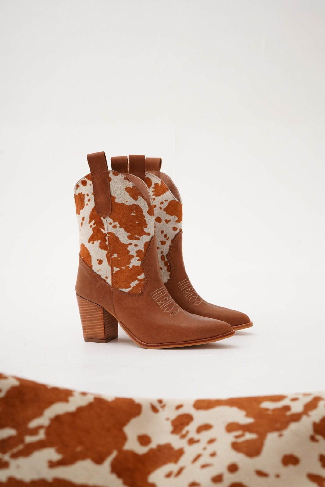 Rich tan leather ankle boots featuring warm orange-and-white cow-print uppers, wooden stacked heels, and engraved detailing. A bold Western statement for stylish day-to-night wear.