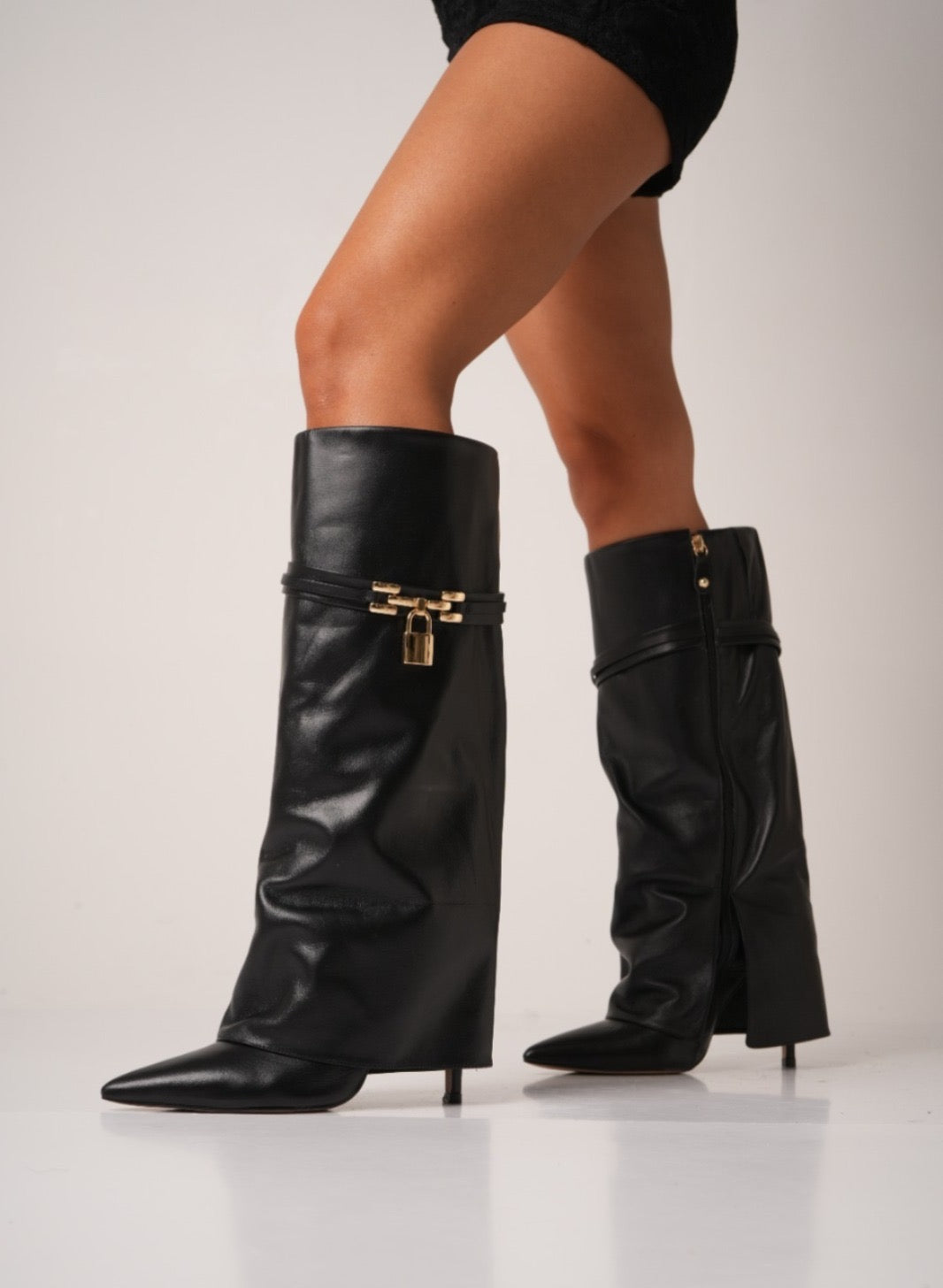Elegant black knee-high stiletto boots with pointed toes, slouchy leather texture, and a gold padlock detail at the ankle. These statement boots blend luxury and edge—perfect for high-fashion looks or a bold night out.