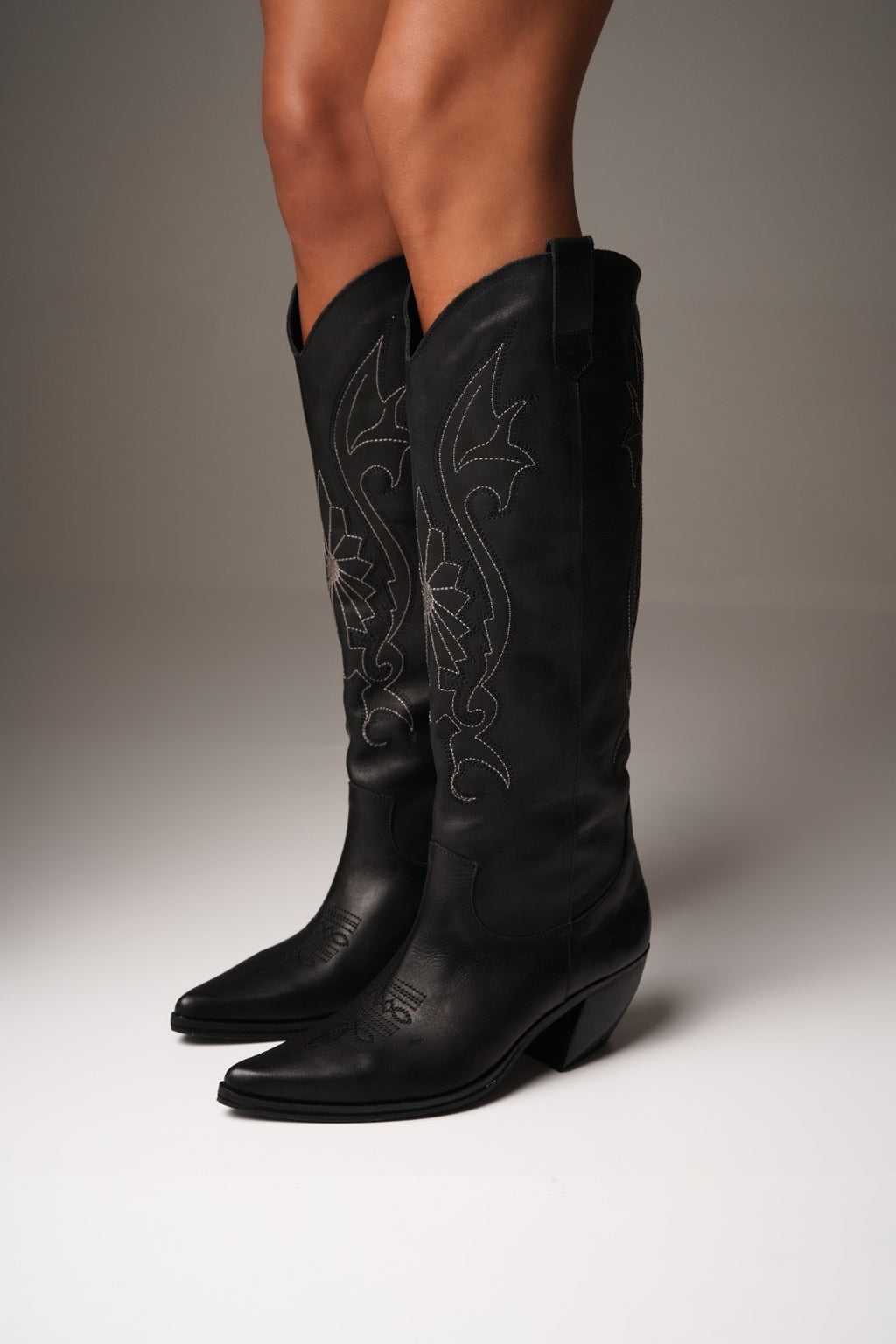 Tall black cowboy boots with pointed toes, angled heels, and subtle black-on-black Western embroidery along the shaft. A sleek and edgy twist on a classic silhouette, perfect for bold, versatile styling.