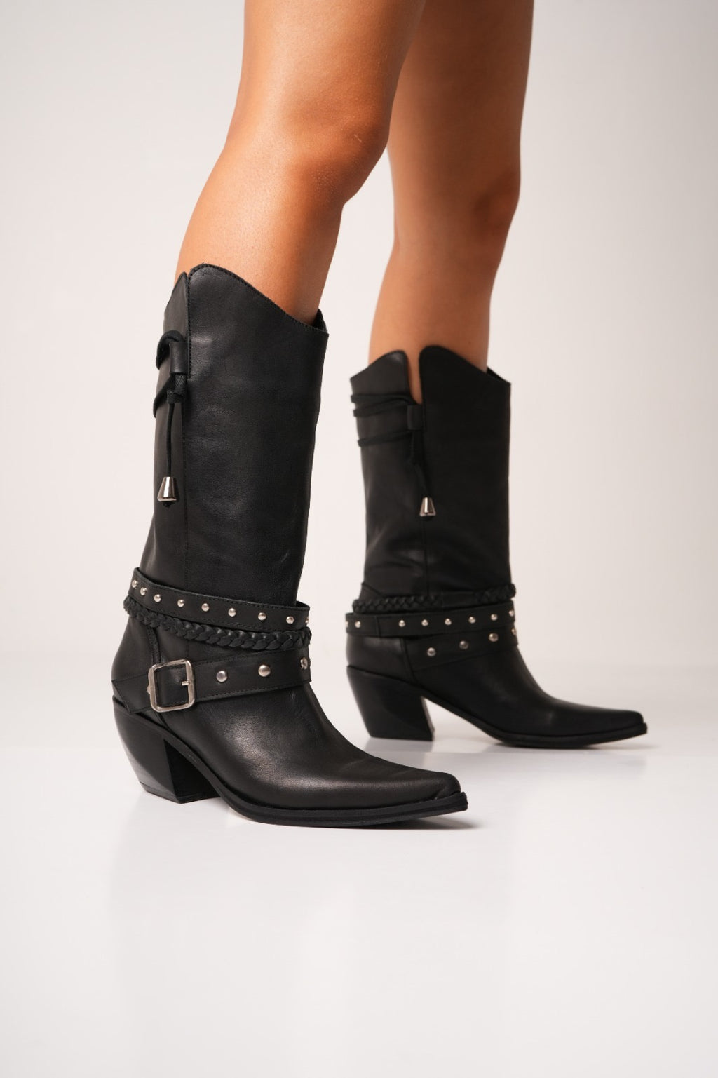Black leather cowgirl boots with mid-height heels, silver studs, braided strap details, and a decorative buckle across the ankle. These sleek western boots feature pointed toes and a modern edge, perfect for bold, fashion-forward styling.