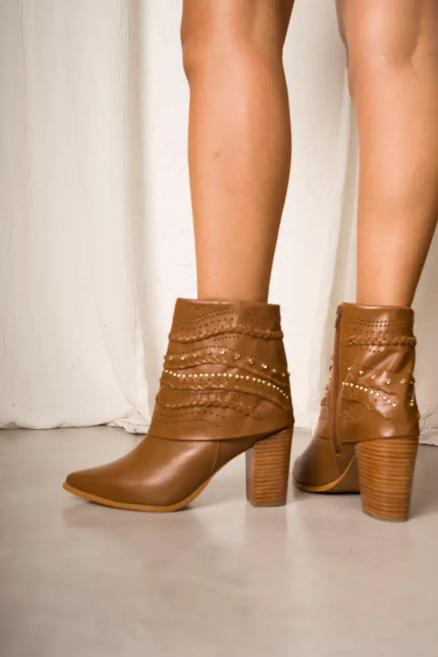 Tan leather ankle boot with a high wooden block heel, featuring braided details, perforated patterns, and small gold studs. A chic Western-inspired design perfect for elevated everyday or boho looks.