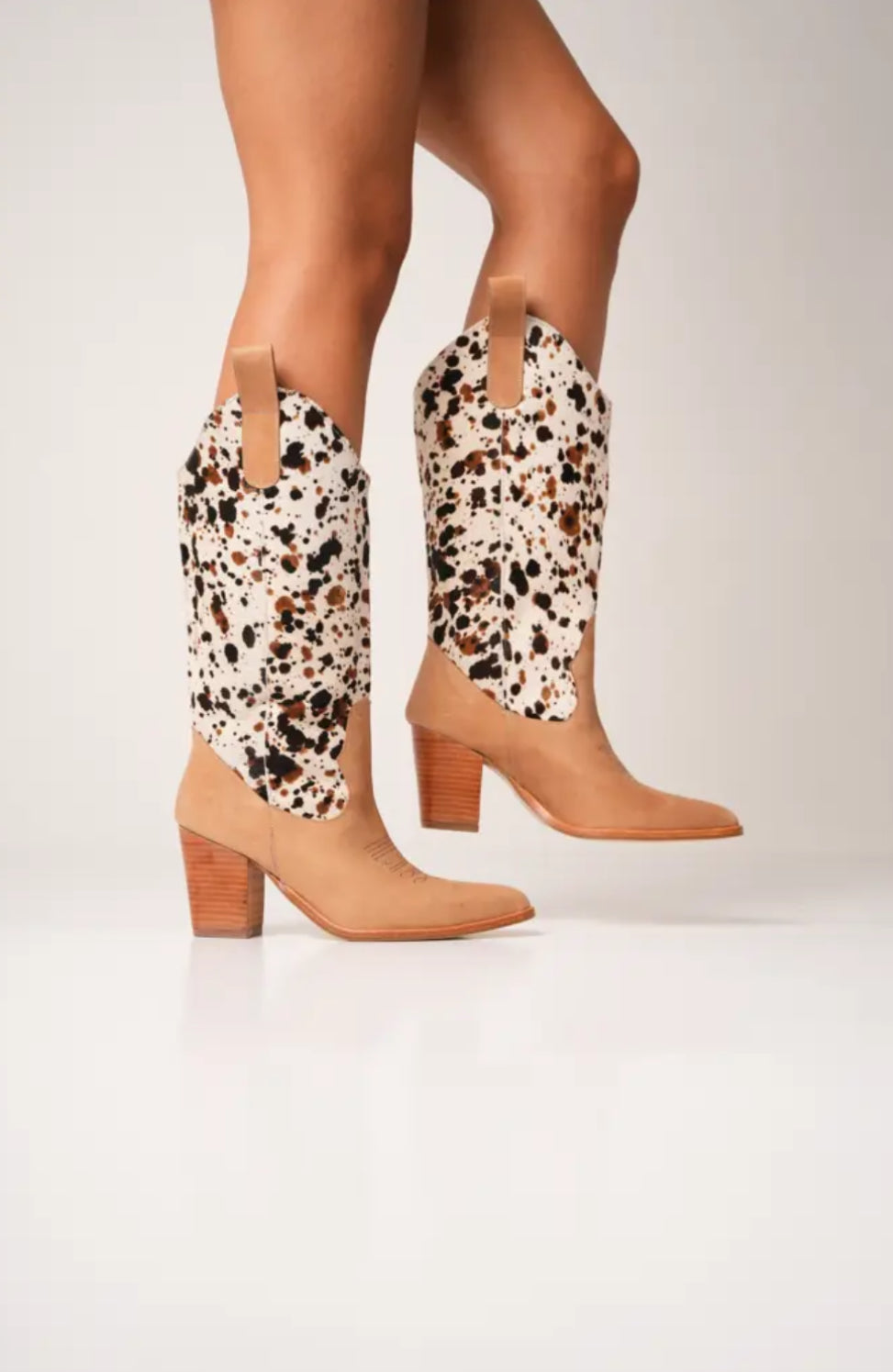 Tan and white cow-print cowboy boots with high block heels, pointed toes, and pull tabs. These bold Western-style boots blend modern flair with ranch-inspired fashion, perfect for standout streetwear looks.