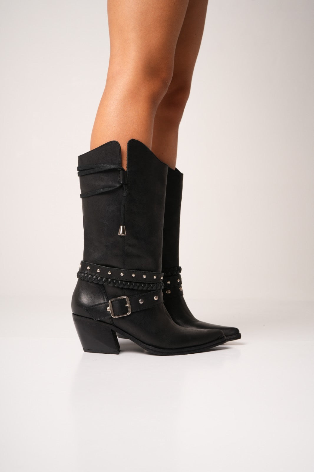 Black leather cowgirl boots with mid-height heels, silver studs, braided strap details, and a decorative buckle across the ankle. These sleek western boots feature pointed toes and a modern edge, perfect for bold, fashion-forward styling.