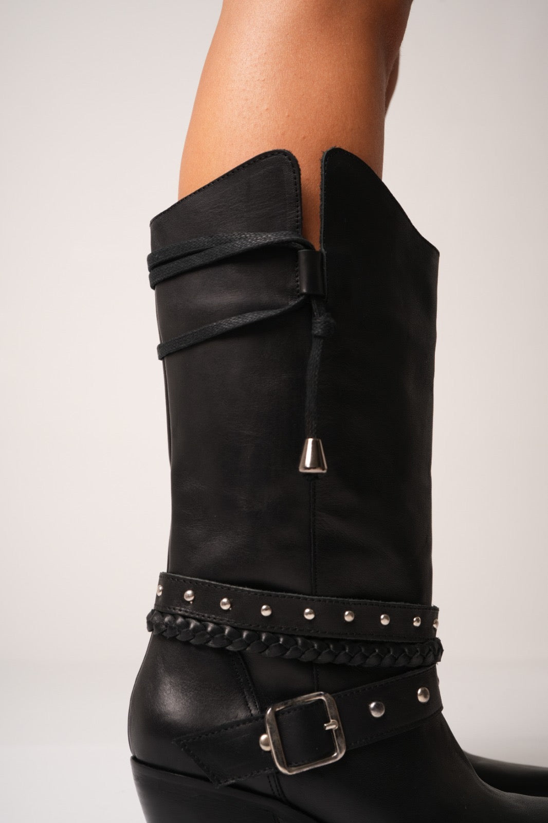 Black leather cowgirl boots with mid-height heels, silver studs, braided strap details, and a decorative buckle across the ankle. These sleek western boots feature pointed toes and a modern edge, perfect for bold, fashion-forward styling.
